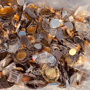 May include: A large collection of assorted metal jewellery findings and pendant trays in various shapes, including oval, round, and rectangular designs. These metallic components feature intricate filigree patterns and smooth finishes in gold, silver, and bronze tones.