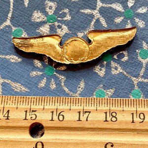 Puede incluir: Un pin de ala de piloto de color dorado con un centro circular. El pin mide aproximadamente 4,5 cm de ancho.