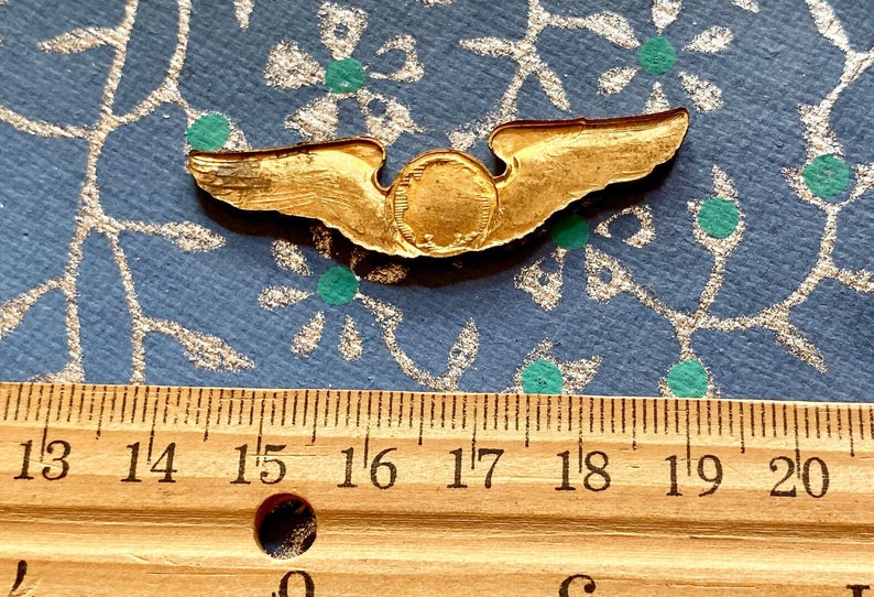 Puede incluir: Un pin de ala de piloto de color dorado con un centro circular. El pin mide aproximadamente 4,5 cm de ancho.
