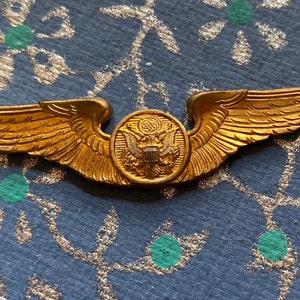 Puede incluir: Una insignia de alas de piloto militar de color dorado con un emblema redondo central que presenta el &aacute;guila americana y el escudo.