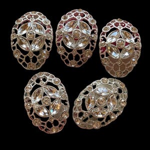 May include: Five oval-shaped decorative buttons featuring a silver-toned metal filigree frame. Each button is embellished with clear, faceted marquise and round crystals arranged in a central geometric pattern, surrounded by a delicate openwork lattice.