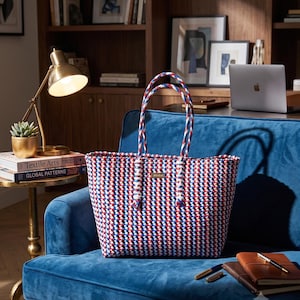 May include: A rectangular tote bag woven from red, white, and blue plastic strips. The bag features two braided handles in matching colors and a small rectangular gold-toned metal plaque on the front center with the text Akidat printed on it.