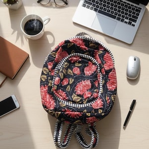 Peut inclure: Un sac à dos noir à imprimé floral avec des fleurs rouges et une bordure rayée blanche est posé sur un bureau en bois. Autres éléments : ordinateur portable, tasse à café, lunettes, stylo, souris, téléphone et petite plante.