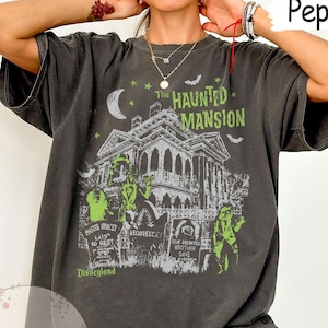 May include: Pepper-colored t-shirt featuring a graphic of The Haunted Mansion, a spooky house with green and white accents, a crescent moon, and bats. The text "The Haunted Mansion" and "Disneyland" are also visible.