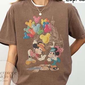 May include: A brown t-shirt featuring a vintage-style graphic of Mickey Mouse and Minnie Mouse holding balloons in front of a castle. The text "Espresso" is in the top right corner of the image.