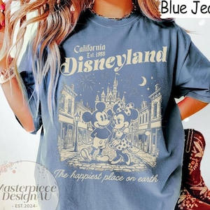 May include: A blue jean coloured t-shirt featuring a cream-coloured graphic of Mickey and Minnie Mouse in front of a castle. The text reads California Est. 1955 Disneyland and The happiest place on earth. The shirt has short sleeves and a crew neckline.