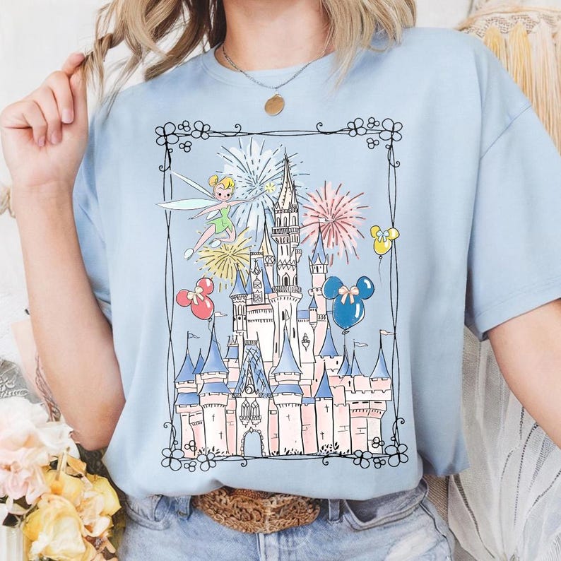 May include: Light blue t-shirt featuring a whimsical illustration of a fairytale castle with fireworks, Tinkerbell, and Mickey Mouse balloons. The design is framed by a black floral border. The shirt is made of a soft material.