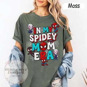 Puede incluir: Camiseta verde musgo con el texto "IN MY SPIDEY MOM ERA" en rojo, blanco y azul. La camiseta presenta personajes de dibujos animados de Spider-Man y telara&ntilde;as. La camiseta es de un material suave.