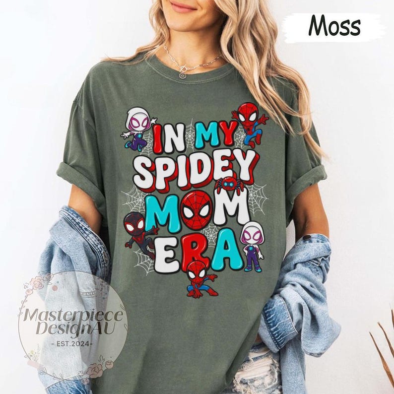 Puede incluir: Camiseta verde musgo con el texto "IN MY SPIDEY MOM ERA" en rojo, blanco y azul. La camiseta presenta personajes de dibujos animados de Spider-Man y telara&ntilde;as. La camiseta es de un material suave.