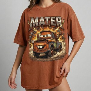 May include: Brown t-shirt featuring a cartoon image of Mater from the Cars movie. The word "MATER" is printed above the character in a stylized font. The graphic includes lightning bolts and tools.