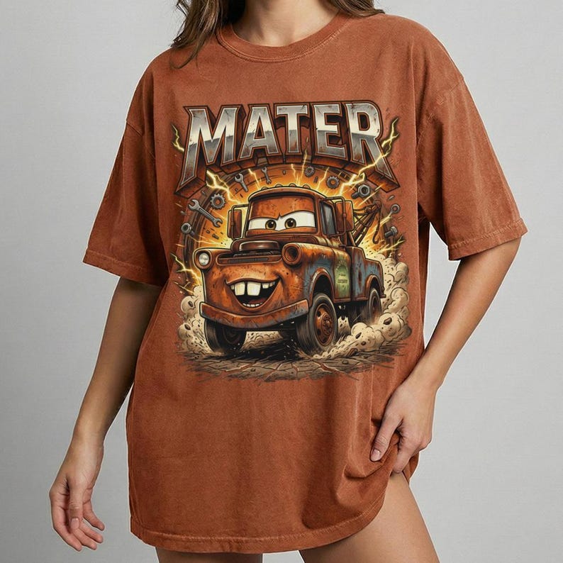 May include: Brown t-shirt featuring a cartoon image of Mater from the Cars movie. The word "MATER" is printed above the character in a stylized font. The graphic includes lightning bolts and tools.