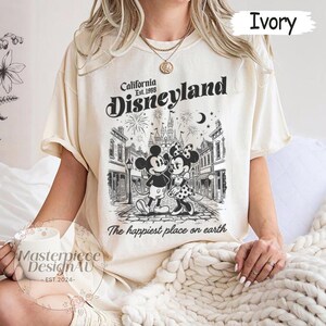 May include: An ivory short-sleeve t-shirt featuring a black graphic print of Mickey and Minnie Mouse in front of a castle. The text reads California Est. 1955 Disneyland and The happiest place on earth. The shirt has a crew neckline and a relaxed fit.