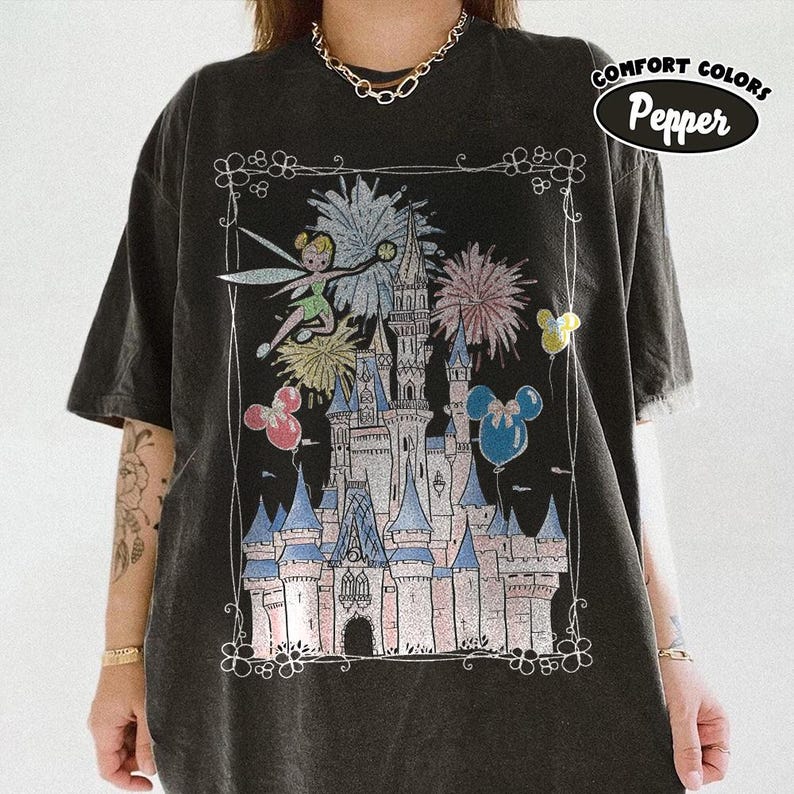 May include: Dark gray Comfort Colors t-shirt featuring a colorful graphic of a fairytale castle, fireworks, Tinkerbell, and Mickey Mouse balloons. The design is framed with a decorative border. The text "Comfort Colors Pepper" is printed on the upper right.
