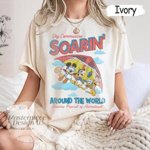 May include: Ivory-colored t-shirt featuring a vintage-style graphic of Mickey Mouse, Donald Duck, and Goofy in a hang glider. The shirt has the text "Sky Commander Soarin' Around The World". The shirt is a relaxed fit.