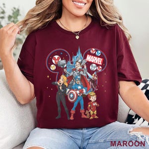 Może przedstawiać: Bordowa koszulka z motywem Marvel inspirowanym Disneyem. Grafika przedstawia postacie Marvela, takie jak Kapitan Ameryka, Thor i Iron Man, na tle zamku. Słowo "MARVEL" jest wyraźnie widoczne.