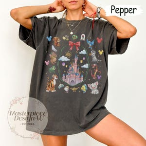May include: Dark gray t-shirt featuring a colorful illustration of a castle surrounded by cartoon characters. The design includes a red bow, clouds, and a sun. The text "Masterpiece DesignAU - EST.2024-" is visible.