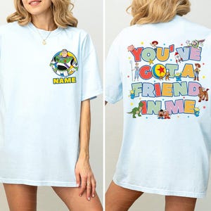 May include: A light blue t-shirt featuring a circular graphic of Buzz Lightyear and the word NAME on the front left chest. The back displays a large colourful graphic with the text YOU'VE GOT A FRIEND IN ME surrounded by various Toy Story characters.