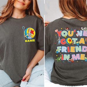 May include: A charcoal grey t-shirt featuring a small graphic on the front chest of Bo Peep with a ball and the text NAME. The back displays the text YOU'VE GOT A FRIEND IN ME surrounded by various Toy Story character illustrations.