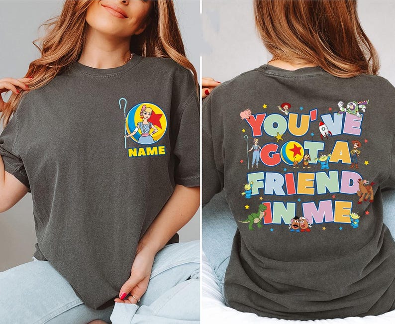 May include: A charcoal grey t-shirt featuring a small graphic on the front chest of Bo Peep with a ball and the text NAME. The back displays the text YOU'VE GOT A FRIEND IN ME surrounded by various Toy Story character illustrations.