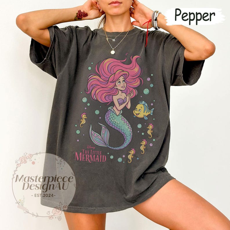 May include: A charcoal gray short-sleeve t-shirt featuring a graphic of a mermaid with long pink hair, a green tail, and a small yellow fish. The text reads The Little Mermaid. The shirt has a crew neckline and a loose, relaxed fit design.