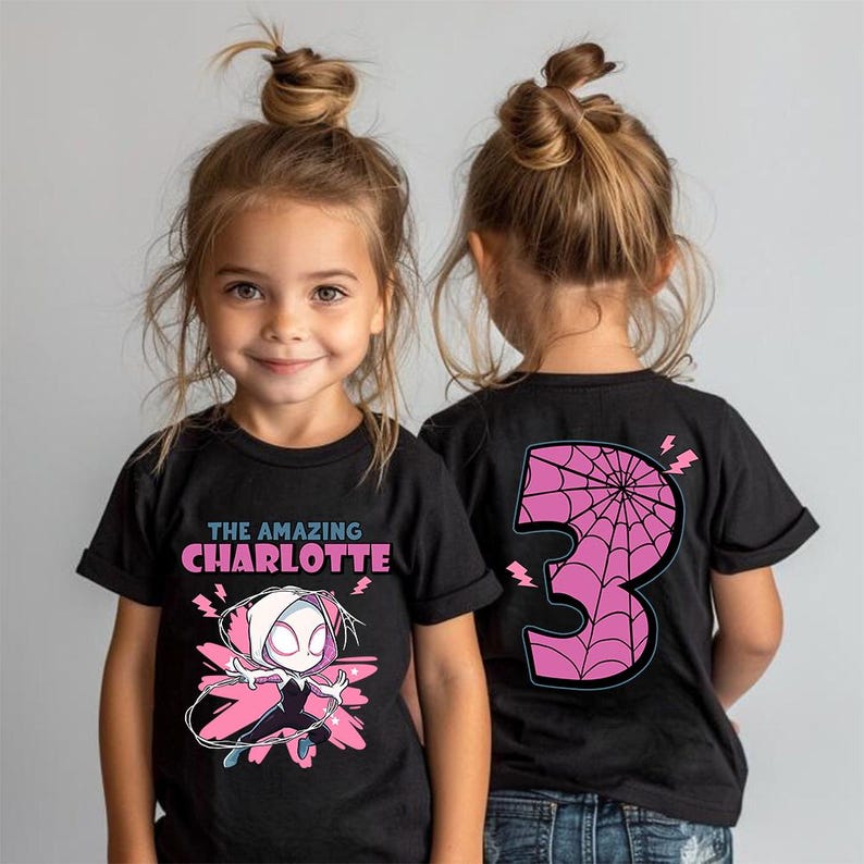 Pu&ograve; includere: T-shirt nere con una grafica di una supereroina e la scritta "THE AMAZING CHARLOTTE" sul davanti. Il retro della maglietta presenta un grande numero 3 rosa con un disegno a ragnatela. Le magliette sono indossate da bambini piccoli.
