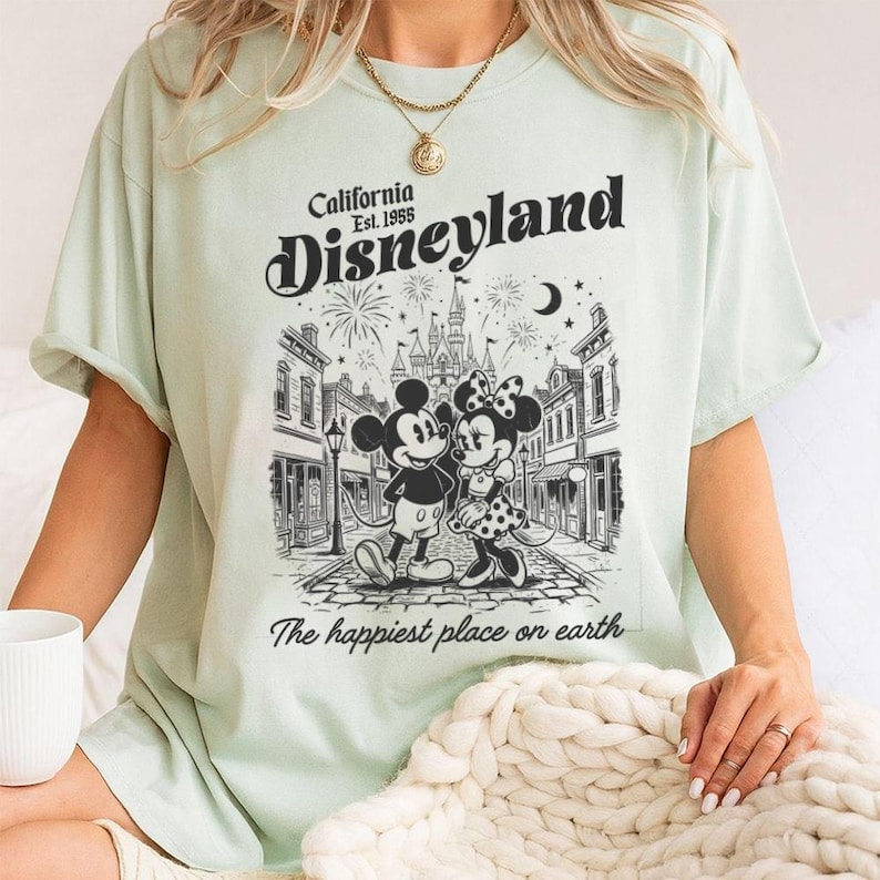 May include: A light green t-shirt featuring a black graphic print of Mickey and Minnie Mouse standing on a street in front of a castle. The text reads California Est. 1955 Disneyland and The happiest place on earth with fireworks and a crescent moon.