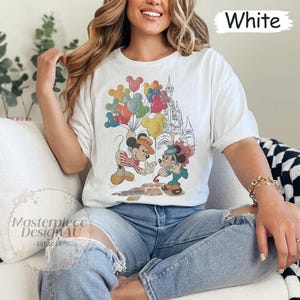 May include: White t-shirt featuring a graphic of Mickey Mouse and Minnie Mouse holding balloons in front of Cinderella's Castle. The text "White" is in the top right corner of the image.