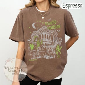 May include: Espresso-colored t-shirt featuring a graphic of The Haunted Mansion, with green and white accents. The design includes the text "The Haunted Mansion" and "Disneyland", along with other themed elements. The shirt is a relaxed fit.