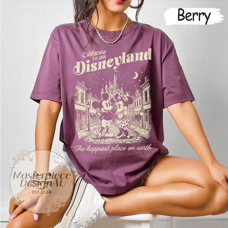 May include: A berry-coloured short-sleeve t-shirt featuring a cream-coloured graphic of Mickey and Minnie Mouse standing on a street in front of a castle. The text reads California Est. 1955 Disneyland and The happiest place on earth.