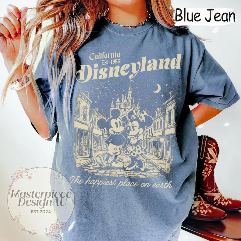 May include: A blue jean coloured t-shirt featuring a cream-coloured graphic of Mickey and Minnie Mouse in front of a castle. The text reads California Est. 1955 Disneyland and The happiest place on earth. The shirt has short sleeves and a crew neckline.