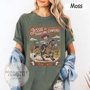 May include: A moss green t-shirt featuring a graphic print of Jessie the Yodeling Cowgirl riding a brown horse in a desert landscape. The design includes the text Jessie the Yodeling Cowgirl, cacti, and two cow skulls at the bottom corners.