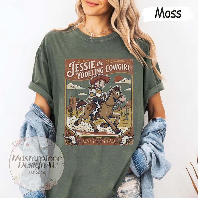 May include: A moss green t-shirt featuring a graphic print of Jessie the Yodeling Cowgirl riding a brown horse in a desert landscape. The design includes the text Jessie the Yodeling Cowgirl, cacti, and two cow skulls at the bottom corners.