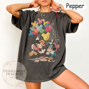 May include: A dark grey t-shirt featuring a vintage-style graphic of Mickey Mouse and Minnie Mouse holding balloons in front of a castle. The text "Pepper" is visible in the top right corner of the image.