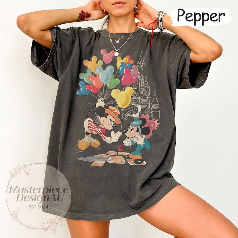 May include: A dark grey t-shirt featuring a vintage-style graphic of Mickey Mouse and Minnie Mouse holding balloons in front of a castle. The text "Pepper" is visible in the top right corner of the image.