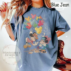 May include: A blue jean t-shirt featuring a vintage-style graphic of Mickey Mouse and Minnie Mouse holding balloons in front of a cartoon castle. The text "Blue Jean" is printed in white on the top right corner of the shirt.