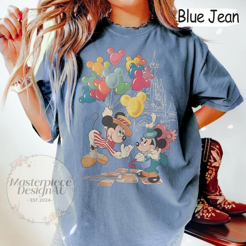 May include: A blue jean t-shirt featuring a vintage-style graphic of Mickey Mouse and Minnie Mouse holding balloons in front of a cartoon castle. The text "Blue Jean" is printed in white on the top right corner of the shirt.