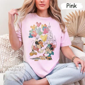 May include: Pink t-shirt featuring a vintage illustration of Mickey Mouse and Minnie Mouse in front of a castle with balloons. The text "Pink" is visible in the top right corner of the image.