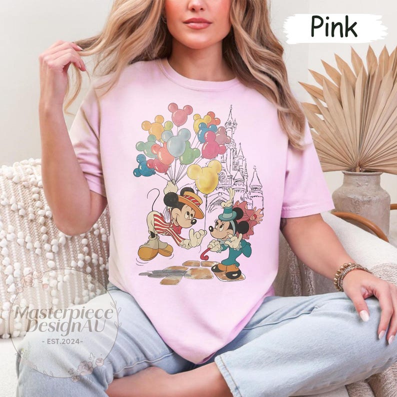 May include: Pink t-shirt featuring a vintage illustration of Mickey Mouse and Minnie Mouse in front of a castle with balloons. The text "Pink" is visible in the top right corner of the image.