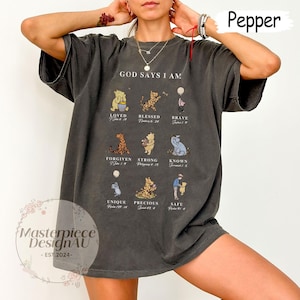 K&ouml;nnte beinhalten: Ein dunkelgraues T-Shirt mit einem Winnie Puuh-Design. Das Shirt zeigt den Text "God Says I Am" und ein Raster mit Winnie Puuh-Charakteren mit Bibelversen.