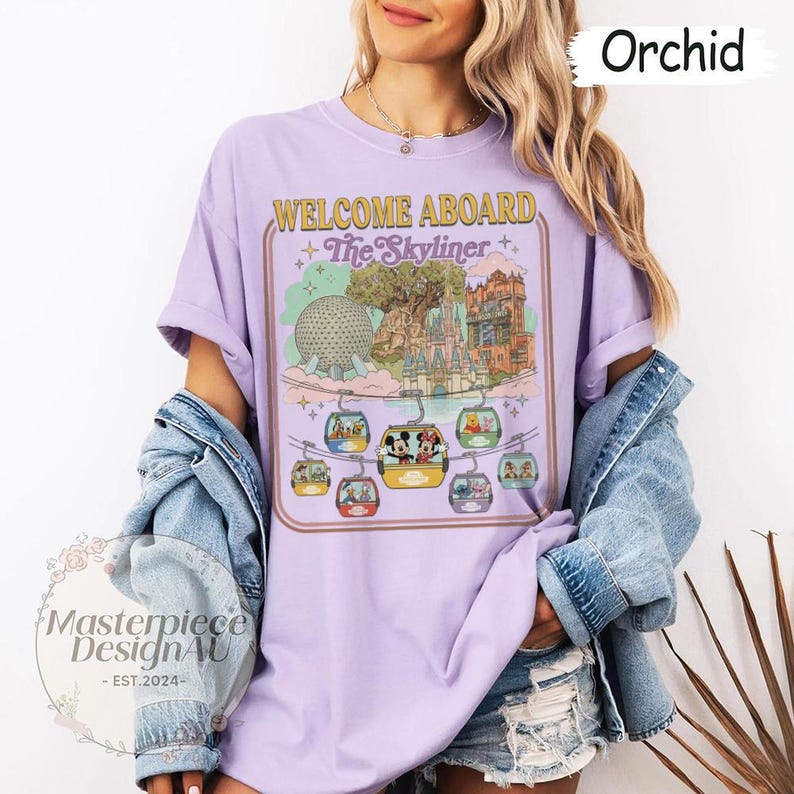 May include: Orchid-colored t-shirt with a graphic that says "WELCOME ABOARD + The Skyliner" featuring a cartoon of a theme park with gondolas. The shirt has short sleeves and a relaxed fit.