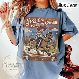 May include: A blue jean colored t-shirt featuring a graphic print of Jessie the Yodeling Cowgirl riding a brown horse in a desert landscape with cacti and mesas. The design includes the text Jessie the Yodeling Cowgirl and two skulls.
