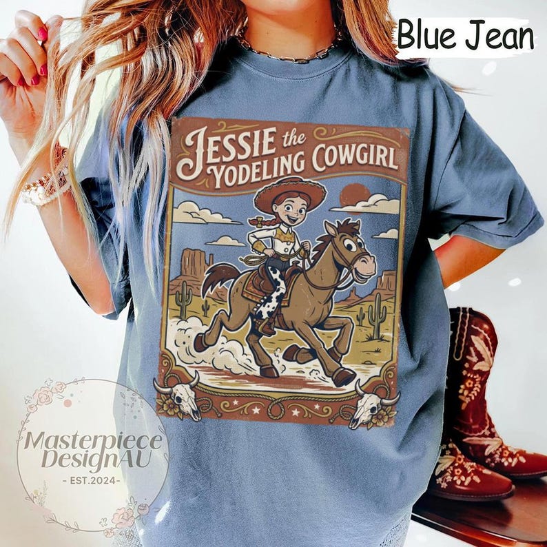 May include: A blue jean colored t-shirt featuring a graphic print of Jessie the Yodeling Cowgirl riding a brown horse in a desert landscape with cacti and mesas. The design includes the text Jessie the Yodeling Cowgirl and two skulls.