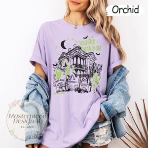 May include: Orchid-colored t-shirt featuring a graphic of The Haunted Mansion, with bats, a crescent moon, and tombstones. The design includes the text "The Haunted Mansion" and "Disneyland". The shirt has rolled-up sleeves.