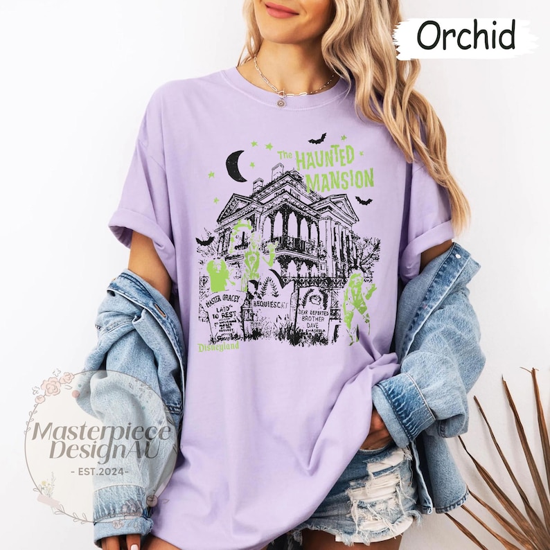 May include: Orchid-colored t-shirt featuring a graphic of The Haunted Mansion, with bats, a crescent moon, and tombstones. The design includes the text "The Haunted Mansion" and "Disneyland". The shirt has rolled-up sleeves.