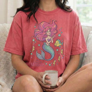 May include: A coral-colored short-sleeve t-shirt featuring a graphic illustration of a mermaid with long pink hair, a green tail, and a yellow fish, surrounded by bubbles and seahorses, with the text Disney The Little Mermaid printed below.