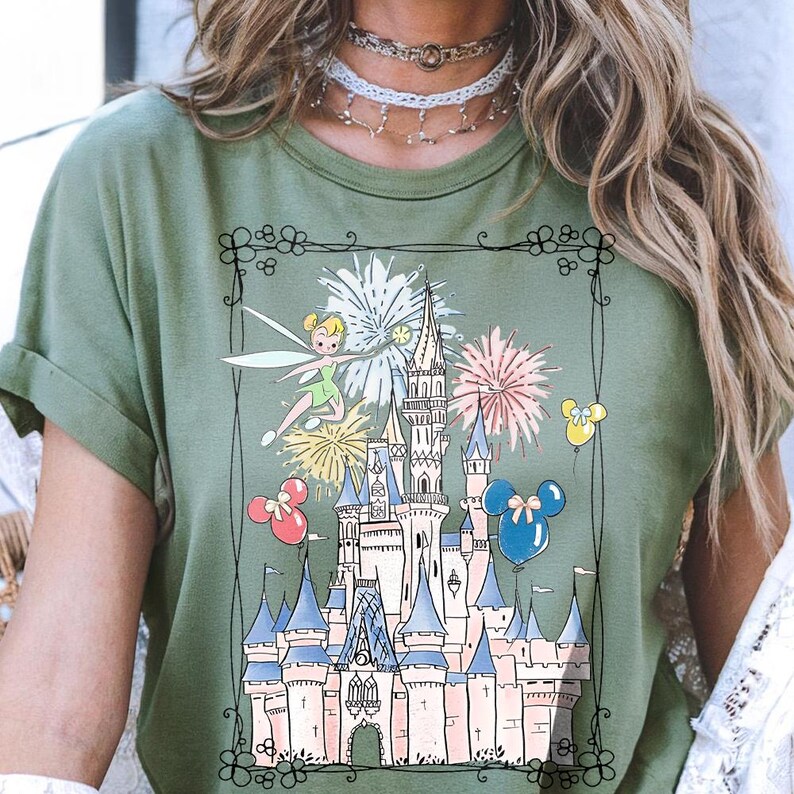 May include: Olive green t-shirt featuring a whimsical illustration of a fairytale castle with fireworks, Tinkerbell, and Mickey Mouse balloons. The design is framed with a decorative black border. The shirt is made of a soft material.