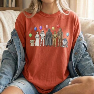May include: A rust-coloured t-shirt featuring a graphic of Star Wars characters, including Baby Yoda, Princess Leia, Darth Vader, and R2-D2, each holding a balloon. The design includes colourful balloons and confetti.