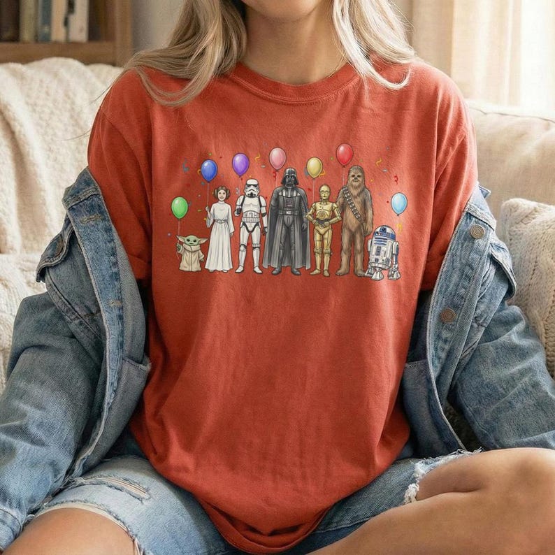 May include: A rust-coloured t-shirt featuring a graphic of Star Wars characters, including Baby Yoda, Princess Leia, Darth Vader, and R2-D2, each holding a balloon. The design includes colourful balloons and confetti.