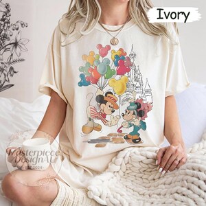 May include: A cream coloured t-shirt with a vintage-style graphic of Mickey Mouse and Minnie Mouse in front of Cinderella's Castle. Mickey is holding a bunch of colourful balloons and Minnie is holding a red rose. The text "Ivory" is in the top right corner of the image.
