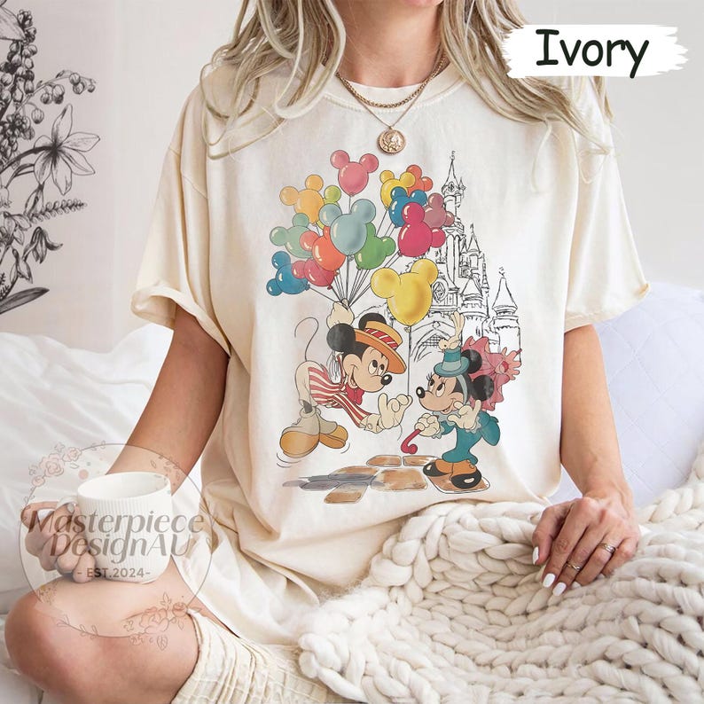 May include: A cream coloured t-shirt with a vintage-style graphic of Mickey Mouse and Minnie Mouse in front of Cinderella's Castle. Mickey is holding a bunch of colourful balloons and Minnie is holding a red rose. The text "Ivory" is in the top right corner of the image.